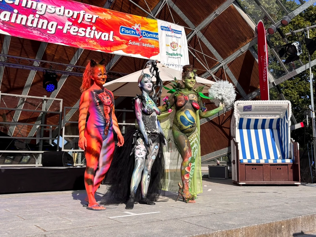 Drei Bodypainting-Models in Orange, Silber und Grün posieren gemeinsam auf der Bühne