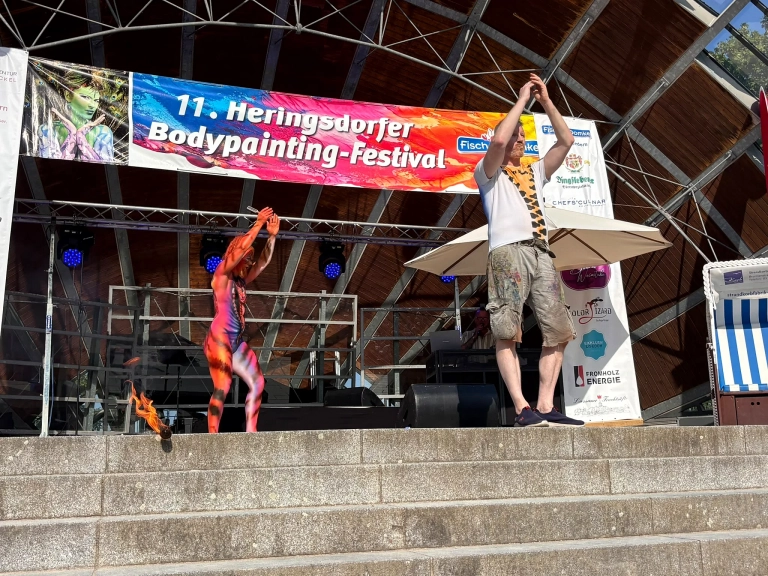 Ein orange bemaltes Model und ein weiterer Performer stehen gemeinsam auf der Festivalbühne