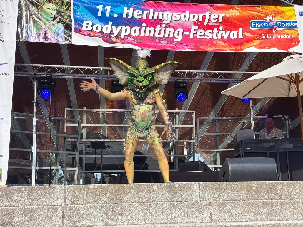 Aufwendig bemaltes Model mit großem grün-braunem Kopfschmuck und ausgestreckten Armen auf der Bühne
