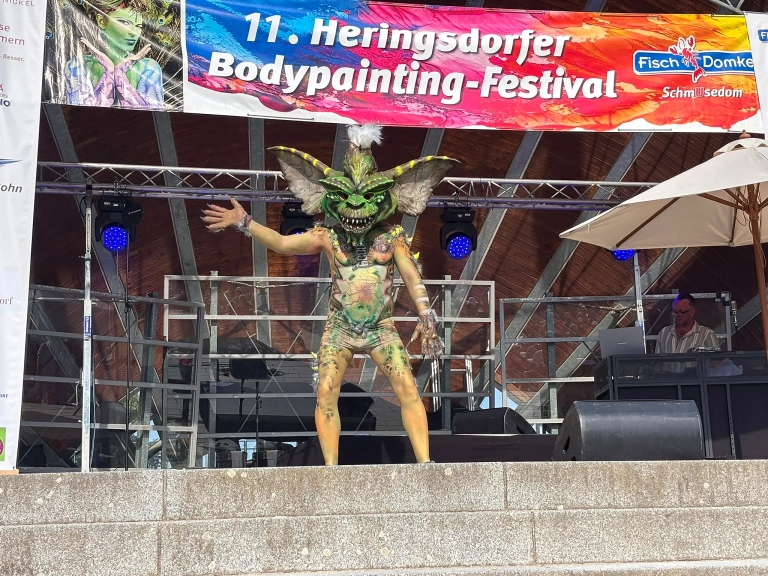 Aufwendig bemaltes Model mit großem grün-braunem Kopfschmuck und ausgestreckten Armen auf der Bühne