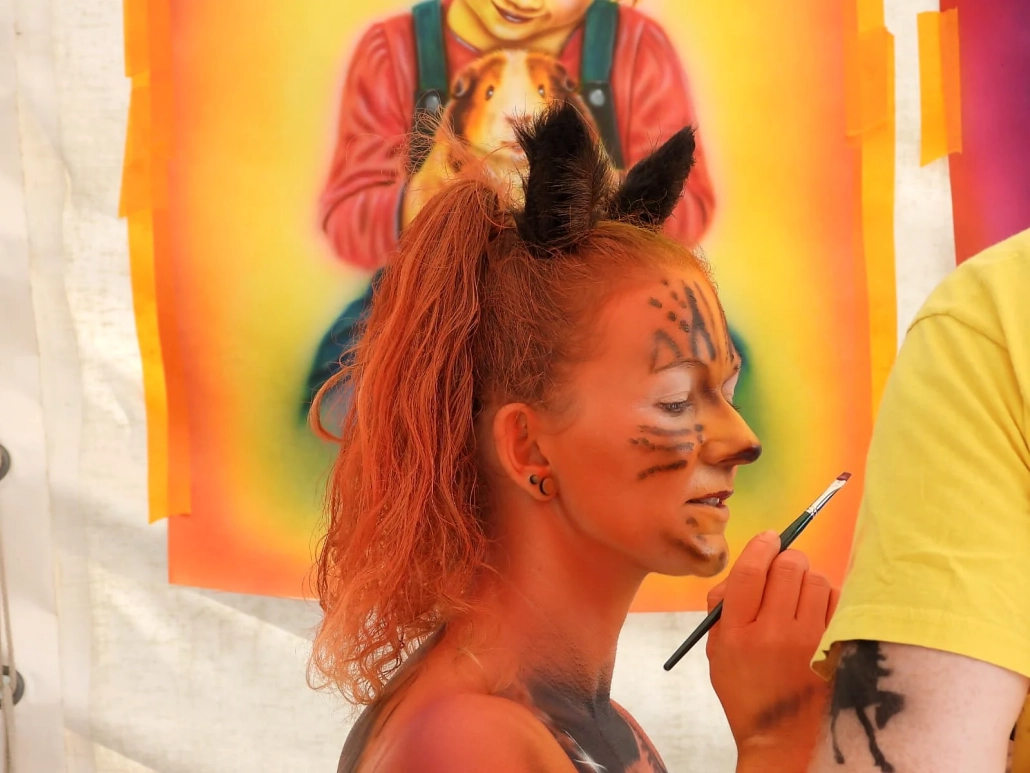 Künstlerin setzt mit einem Pinsel feine Akzente im Gesicht eines orange bemalten Models