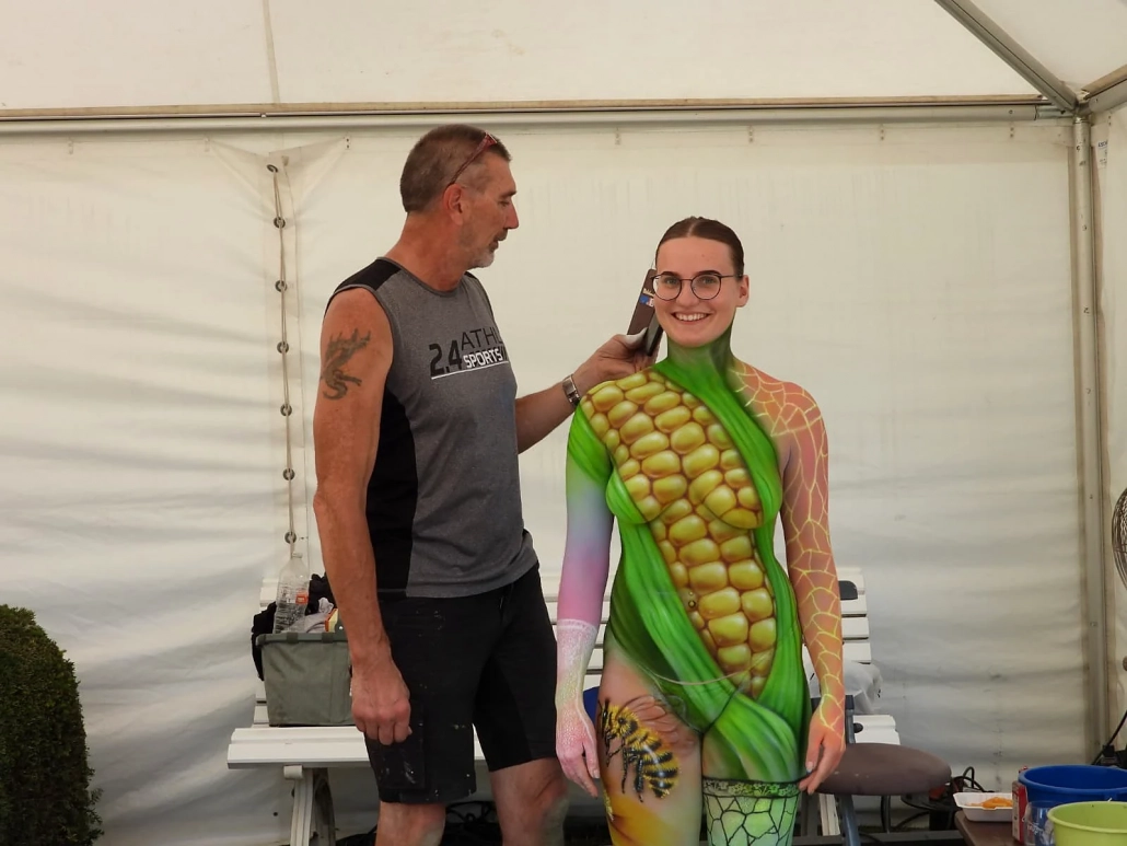 Grün-gelb bemaltes Model mit Maismotiv steht mit einem Begleiter im Festivalzelt