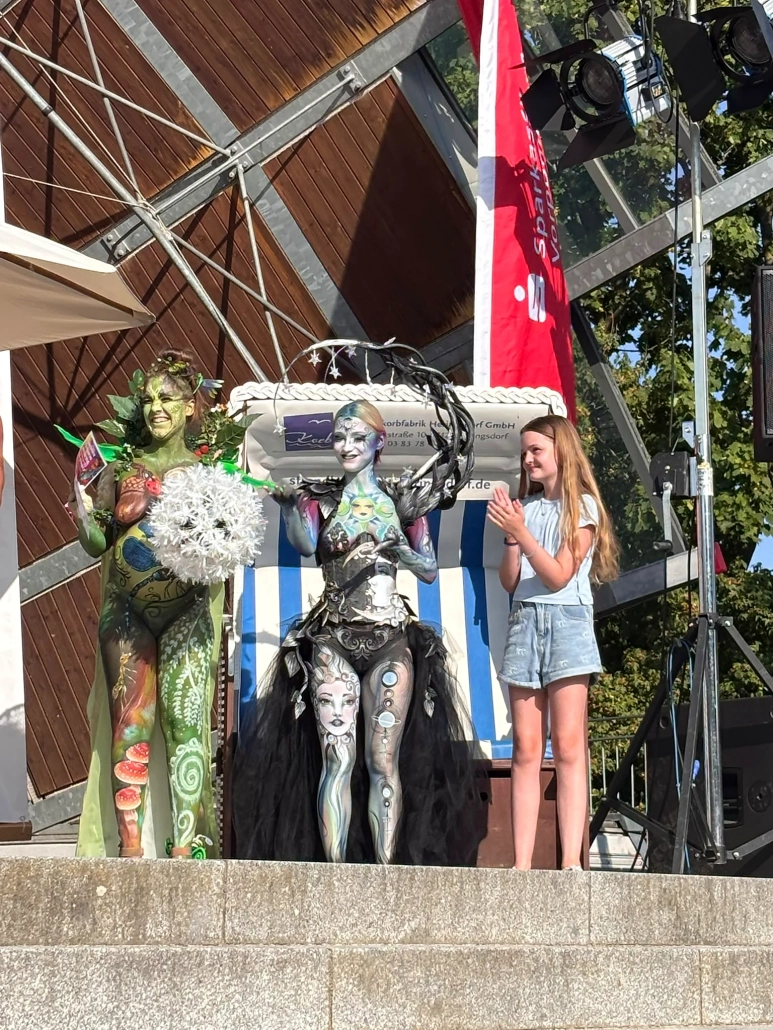 Ein silbernes und ein grünes Bodypainting-Model stehen nebeneinander auf der Bühne