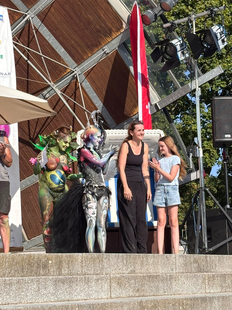 Zwei Bodypainting-Models stehen mit einer weiteren Person am Rand der Festivalbühne