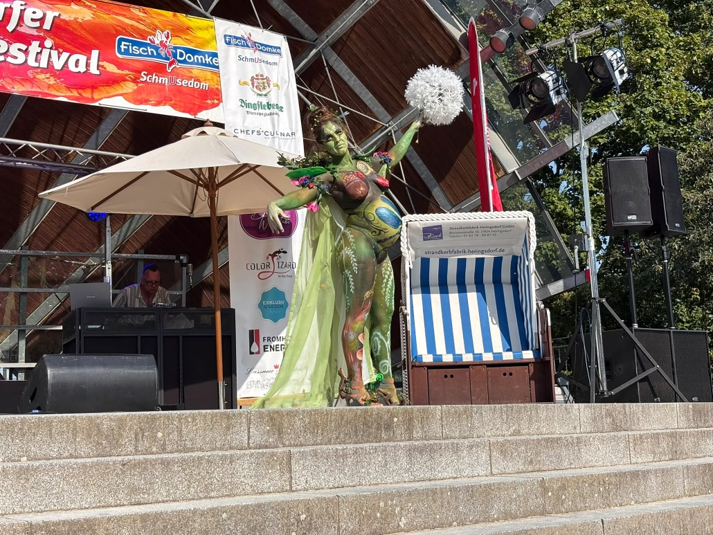 Grün bemaltes Model mit Kopfschmuck und fließendem Kostüm bewegt sich über die Festivalbühne