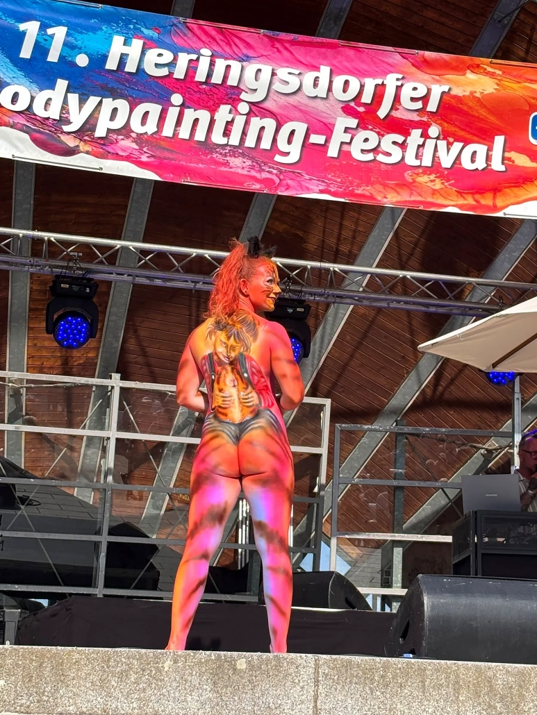 Orange und violett bemaltes Model steht in selbstbewusster Ganzkörperpose auf der Festivalbühne