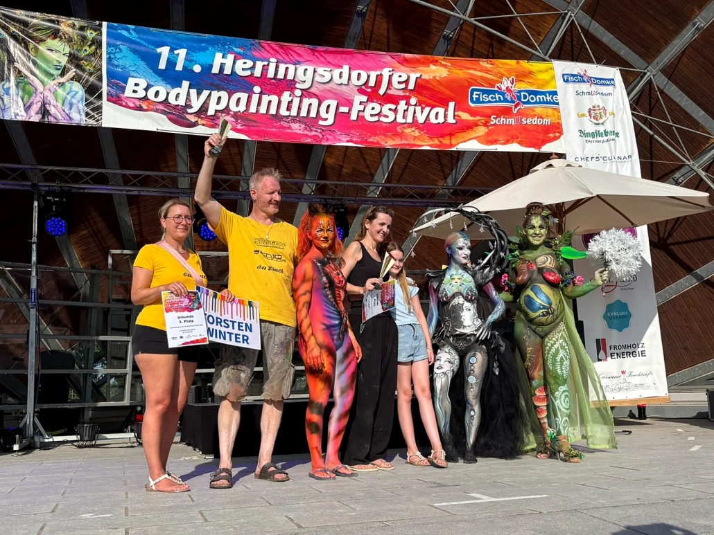 Ausgezeichnete Teilnehmende und mehrere Bodypainting-Models stehen gemeinsam vor dem Festivalbanner