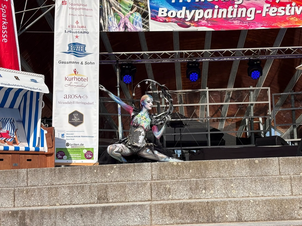 Schwarz-silbern bemaltes Model kniet in einer symmetrischen Pose auf der Festivalbühne