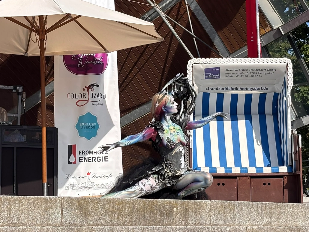 Schwarz-silbern bemaltes Model sitzt in einer dramatischen Pose neben einem Strandkorb auf der Bühne